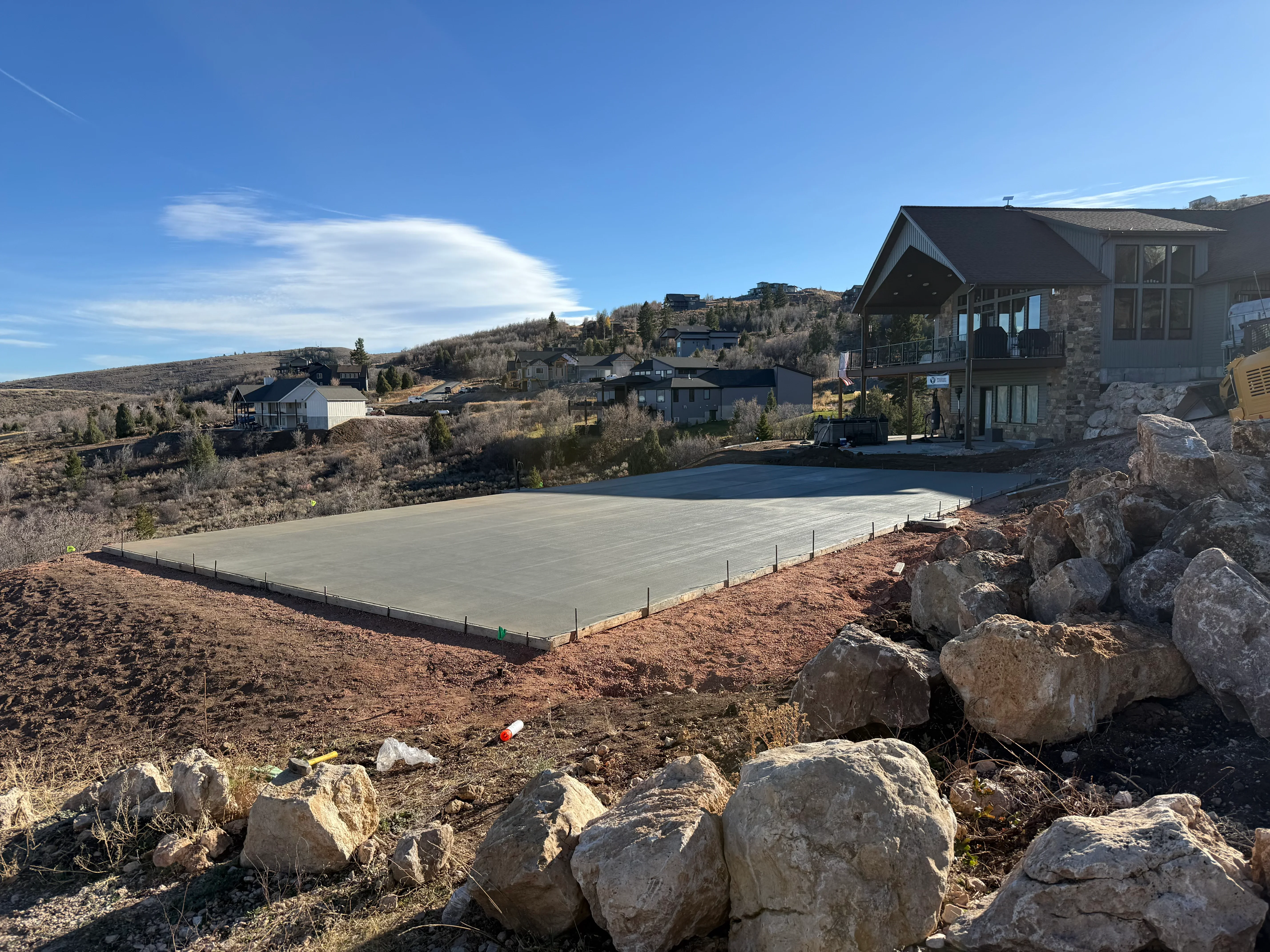 Fresh concrete foundation pad with edge forms on sloped mountain terrain, large boulders visible