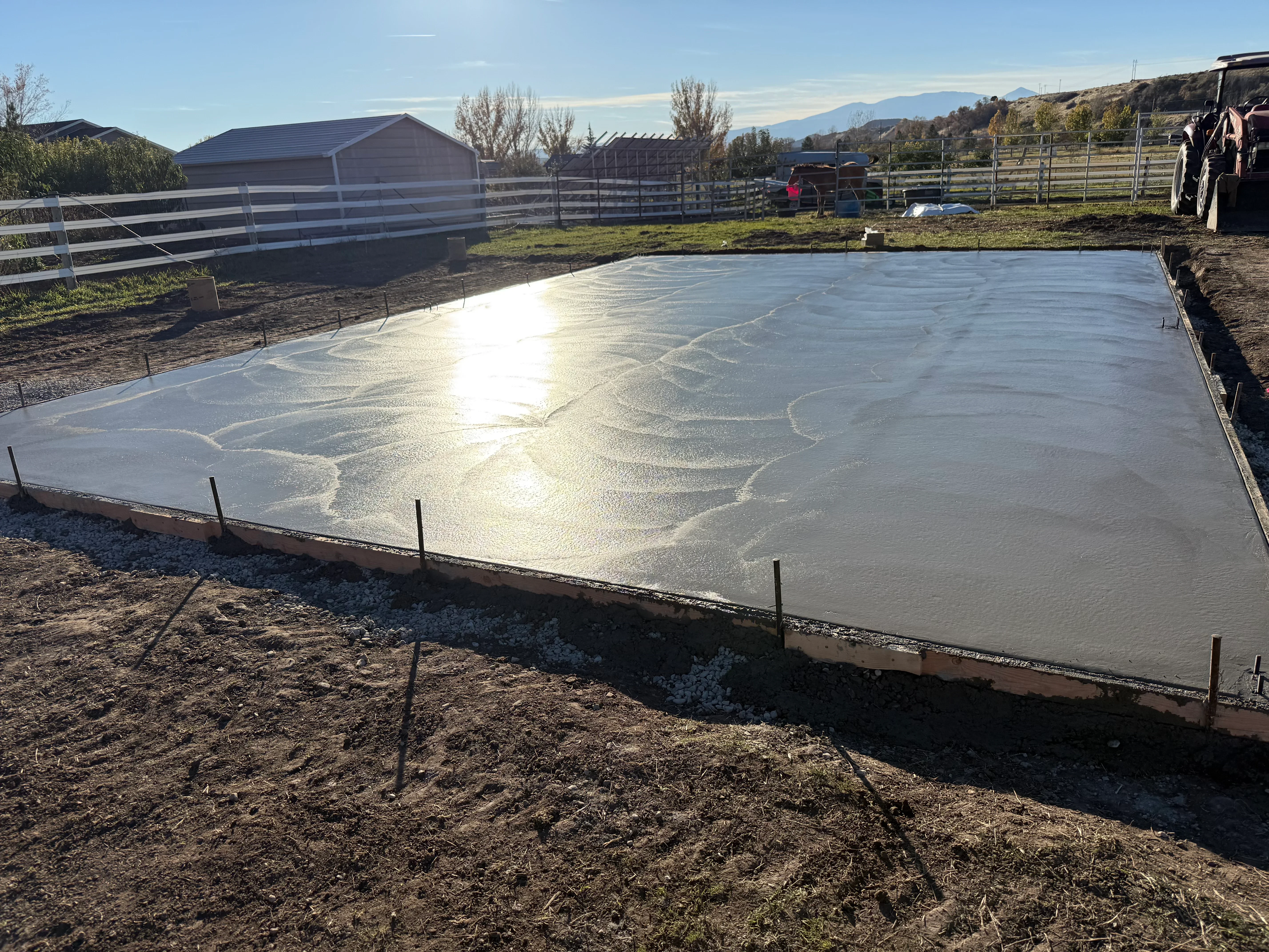 Freshly poured concrete foundation with gravel base, steel rebar stakes, and wooden forms on rural property